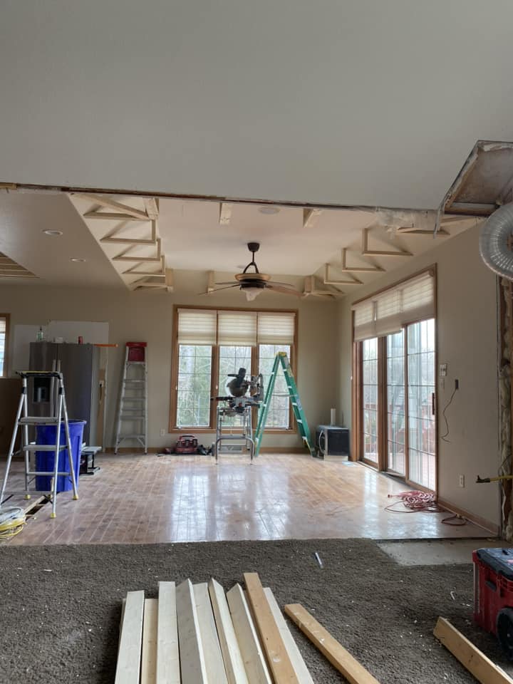 Great room during active renovation with ceiling drywall removed, miter saw on scaffolding, and lumber staged on floor